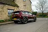 Mørkerød Kia EV5 parkert ved en steinbygning langs en smal landevei.